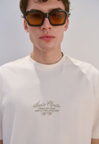 T-shirt beige di cotone con testo ricamato "Santa Clarita" in marrone chiaro, abbinata a occhiali da sole neri oversize con lente ambrata.