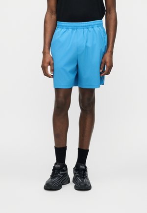 Mężczyzna w jasnoniebieskich spodenkach z elastycznym pasem, czarnych skarpetach do kostek i czarnych sneakersach Diesel, stoi na tle jednolitego białego tła.