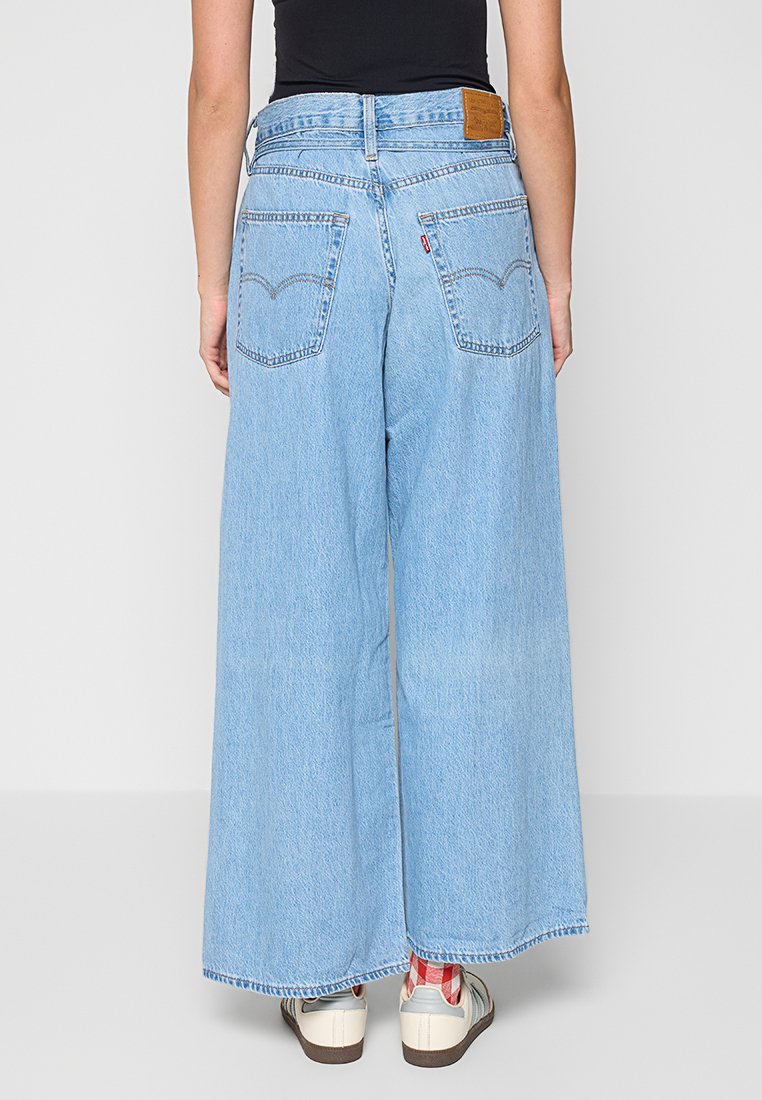 Un jean large bleu clair avec une taille haute, deux poches arrière et une étiquette en cuir beige au niveau de la ceinture arrière. Matériau : denim.