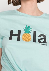 Jasnoniebieska bawełniana koszulka z czarnym napisem "Hola" i żółtą grafiką ananasa, z napisem "TROPICAL" na dole.