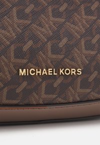 Καφέ τσάντα με σ χέδιο που φέρει το χρυσό λογότυπο "MICHAEL KORS". Το υλικό έχει υφή που μοιάζει με υφαντό και διαθέτει σταθερή καφέ βάση.