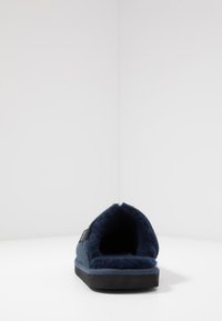 Blå filt hjemmesko med blødt pelsfor, åben bagside, robust sort sål og en tekstureret overflade. Minimalistisk design med en diskret logoskilt.