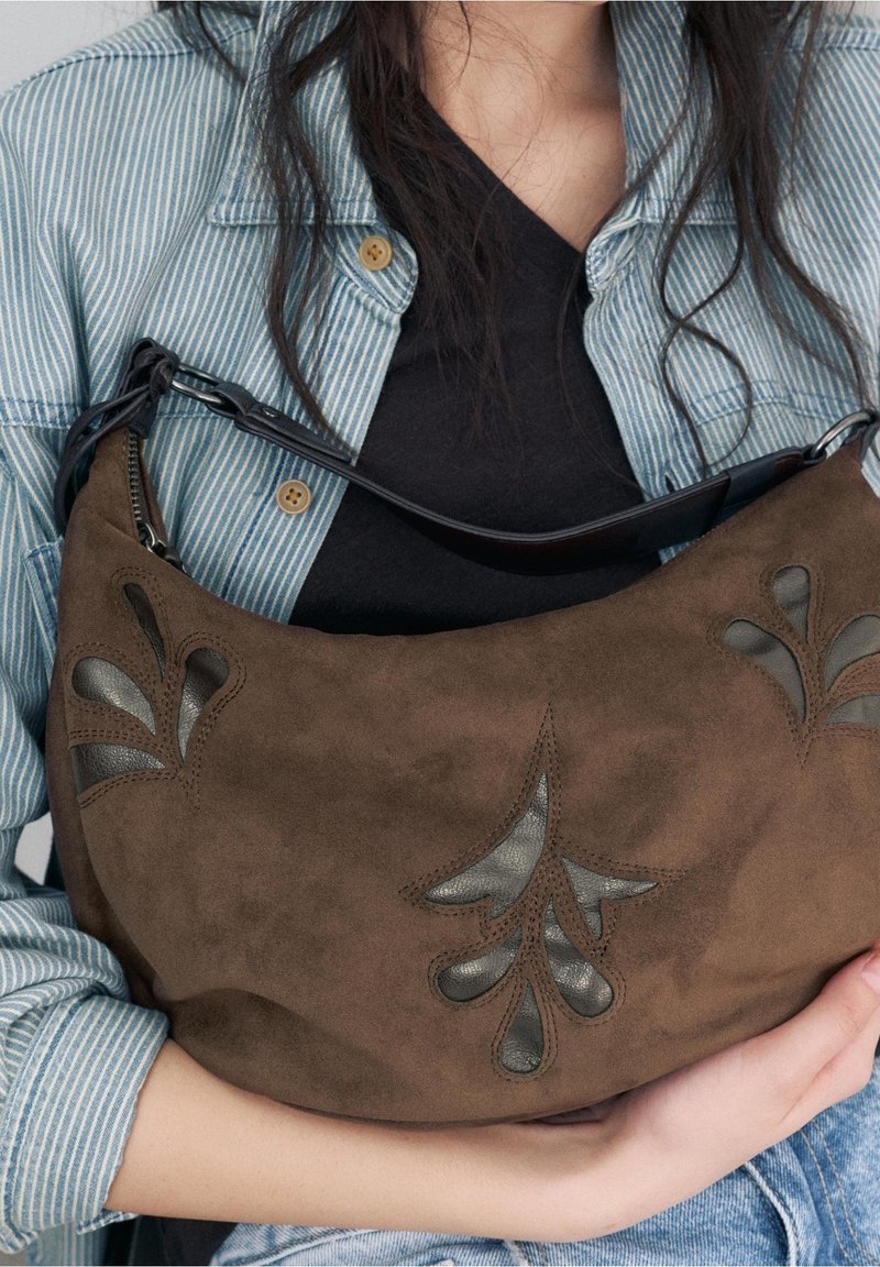 Femme portant une chemise rayée tient un sac à main en daim marron avec des découpes florales en cuir noir sur le devant.