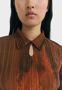 Bruine geplooide blouse met een sleutelgat-halslijn, een strakke kraag en subtiele textuurvariaties; bevat verticale strepen en een lichte glans.