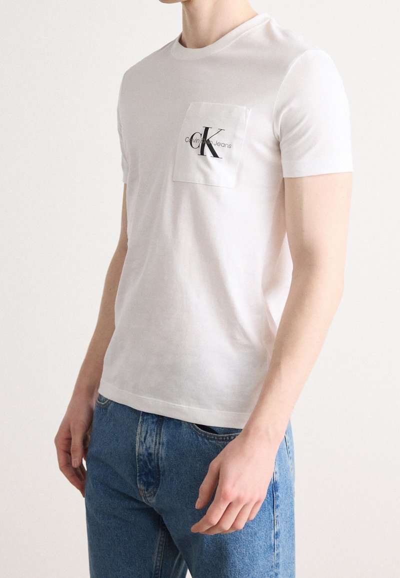 Muž v bílém tričku Calvin Klein Jeans s logem na kapsičce a modrých džínách, stojící na pozadí s jemným světlým odstínem.