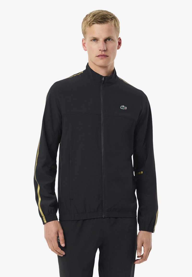 Junger Mann trägt eine schwarze Sportjacke mit Reißverschluss, die goldene Streifen an den Ärmeln und ein kleines grünes Krokodil-Logo auf der Brust hat.