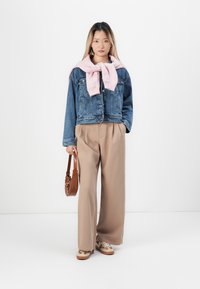Denim jacket with a pink sweater tied around the neck, tan wide-leg trousers, and patterned shoes. Brown shoulder bag completes the outfit.