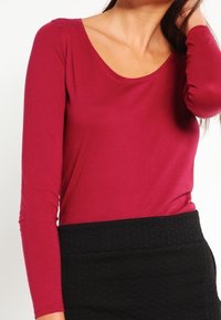 Långärmad bordeaux top med rundad halsringning, tillverkad av mjukt tyg. Designad med en figurnära silhuett och en strukturerad svart kjol.