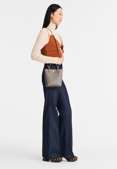 Braun Wildlederweste mit zwei Vorderaschen, cremefarbener gerippter Rollkragenpullover, hochtaillierte dunkelblaue Jeans und eine kleine graue Handtasche. Schuhe mit Holzabsatz.
