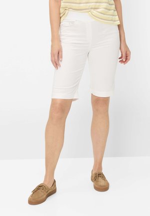 Kvinde iført hvide knælange shorts, en gulstribet top og tanfarvede snørebånds-casualsko, stående mod en ensfarvet baggrund.