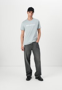 Világoskék pamut póló "BELSTAFF" felirattal fehér színben, laza szabású sötétszürke farmernadrággal és fekete cipővel, sima háttér előtt.