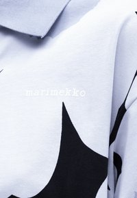 Svetlomodrá látka s veľkými čiernymi abstraktnými tvarmi a bielym textovým logom "marimekko" vyšívaným blízko okraja goliera.