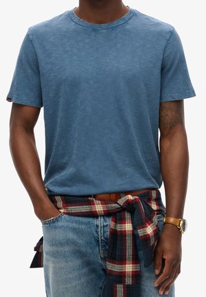 Blaues T-Shirt mit Rundhalsausschnitt aus weichem Stoff, getragen mit einem rot-navy-karierten Flanellhemd, das in der Taille über hellblauen Jeans gebunden ist.