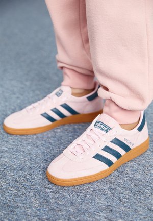 Persona con zapatillas Adidas Spezial rosas con suelas de goma y rayas azul oscuro, de pie sobre un suelo alfombrado azul.