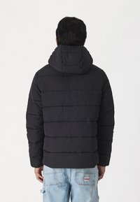 Svart vadderad jacka med huva, med horisontella quiltade sektioner och en slät yta. Designen är enkel och funktionell.