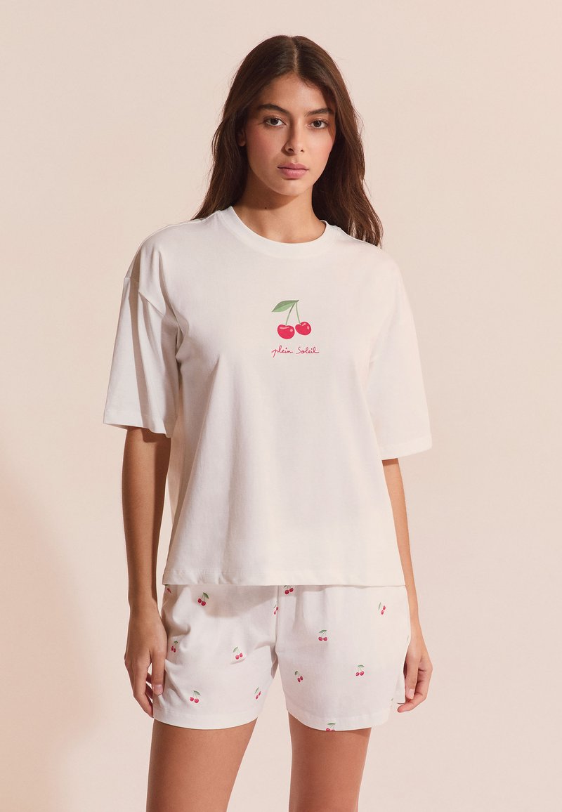 Femme portant un t-shirt blanc à manches courtes avec un motif de cerise et un short blanc assorti avec de petites impressions de cerises, debout devant un fond beige.