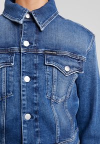 Detailní záběr na modrou džínovou bundu Calvin Klein se stříbrnými logovými knoflíky a náprsními kapsami na osobě.