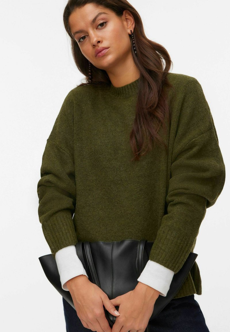 Pull overdimensionné vert foncé avec des poignets et un col côtelés, associé à une pochette noire. Le tissu semble doux et texturé.