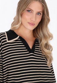 Maglione a strisce nere e beige con scollo a V e colletto a contrasto. Tessuto morbido, vestibilità rilassata e strisce distintive creano un interesse visivo.