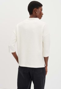 Persona con capelli corti che indossa un top bianco a maniche tre quarti traslucido e pantaloni neri, vista da dietro.
