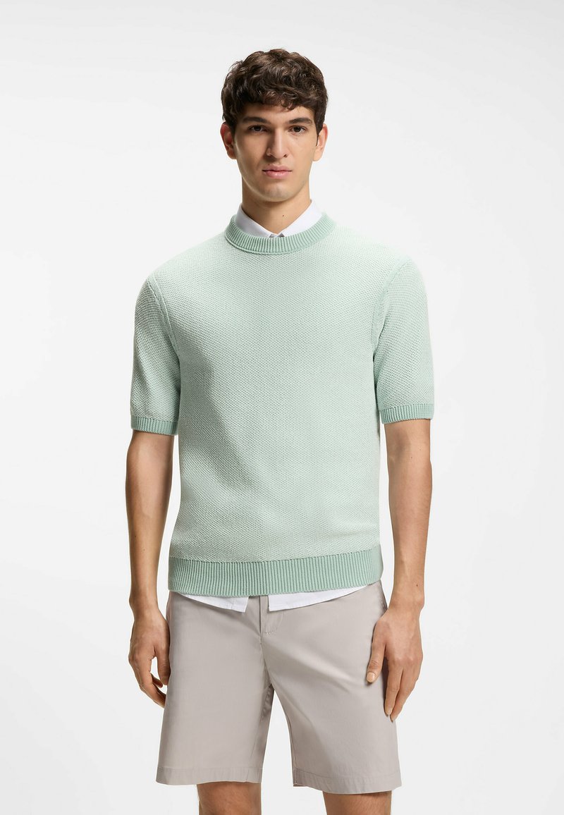 Jeune homme debout portant un pull en tricot à manches courtes vert clair sur une chemise blanche et un short beige au genou, sur un fond blanc.