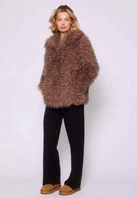 Brun fluffig pälsimitation med bred krage, oversized passform och långa ärmar, kombinerad med svarta byxor och bruna skor.