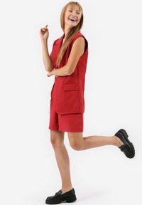Rode tweedelige outfit met een mouwloze blazer en bijpassende short. Het materiaal heeft een gladde textuur. Zwarte schoenen maken de look compleet.
