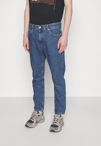 Blå denimjeans med rak skärning, fem fickor, knäppning med knapp och minimal blekning, i kombination med grå och rosa sneakers.