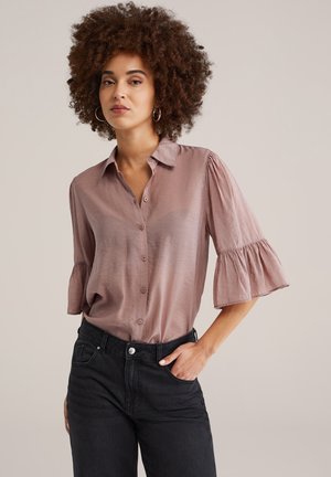 Femeie cu păr creț, purtând o bluză mauve cu nasturi și mâneci jumătate cu volane și blugi negri cu talie înaltă, mâna în buzunar, fundal neutru.