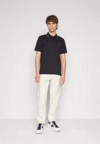 Black polo shirt with white trim, soft cotton material. Paired with light-colored trousers and black and white sneakers. Model stands upright.