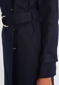 Närbild av en marinblå kappa med ett brett bälte med metallring, sned sidoficka och rem detaljer på ärmen.