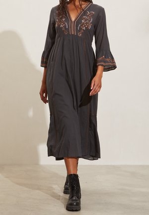 Femme portant une robe midi noire avec broderies florales et manches évasées, associée à des bottes de combat noires à lacets, marchant à l'intérieur.