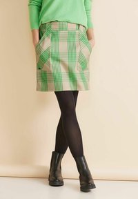 Beige Karorock mit grünen Streifen, vorne mit Taschen und geradem Schnitt. Kombiniert mit schwarzen Strumpfhosen und Stiefeletten.