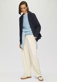 Marineblauer Trenchcoat über einem hellblauen strukturierten Pullover, kombiniert mit cremefarbenen weiten Hosen und weißen Sneakern mit braunen Akzenten.