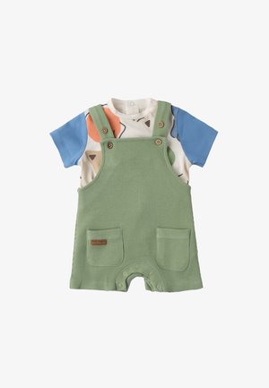 Salopette a quadretti verdi con bottoni di legno, dotata di due tasche frontali. Sotto, una camicia a maniche corte con forme astratte colorate.