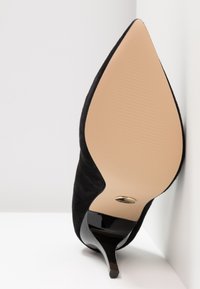 Chaussure à talon haut en daim noir à bout pointu, avec semelle beige et talon aiguille noir brillant, présentée en position verticale sur une surface blanche.