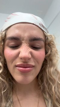 Nahaufnahme einer Person mit lockigem Haar, die ein weißes Kopftuch mit roten Linien trägt und einen Gesichtsausdruck mit verzogenem Gesicht zeigt.