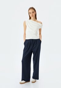 Weißes Top mit einem Schulterträger, kombiniert mit navyblauen, weit geschnittenen Hosen und weißen Sneakers. Glattes Material und lockere Passform mit Seitentaschen.