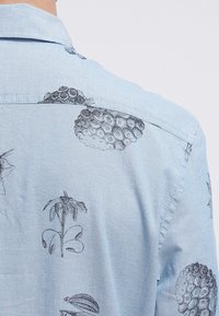 Lichtblauw shirt met zwarte botanische en fruittekeningen, waaronder bananen, bloemen en gegroepeerde cirkelvormige fruitpatronen.
