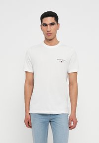 Tricou alb din bumbac cu mâneci scurte și guler rotund, având un mic logo negru în zona stângă a pieptului, asociat cu blugi ușor albaștri.