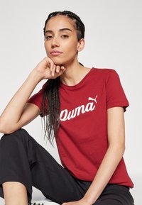 Czerwona bawełniana koszulka z białym logo i emblematem "Puma". Krótkie rękawy, okrągły dekolt i luźny fason. Widać teksturowane ciemne spodnie.