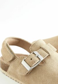 Sandale en suède beige avec une bride réglable et une boucle en métal argenté. Semelle en liège et semelle texturée, dotée d'un design arrondi.