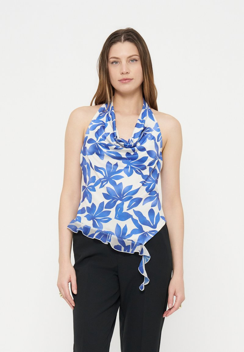 Femme portant un top à col halter bleu et blanc à motif floral avec un ourlet asymétrique, associé à un pantalon noir, debout devant un fond uni.
