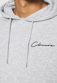 Grijze hoodie met trekkoorden, voorzien van een zwarte cursieve logo op de borst. Het materiaal is zacht en gestructureerd, met een klassieke pasvorm.