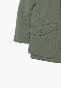 Olivgrüne Parkajacke mit glatter Textur, ausgestattet mit einer großen Aufsetztasche und Druckknopfdetails an der Vorderseite.