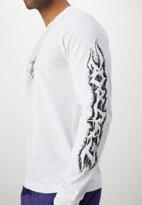 Valkoinen pitkähiha t-paita, jossa oikeassa hiassa musta graafinen kuvio, joka esittelee rohkeita, abstrakteja muotoja ja tekstuureja.