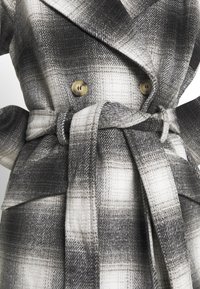 Cappotto doppiopetto a quadri nelle tonalità di nero, grigio e bianco. Presenta bottoni dorati e una cintura abbinata. Tessuto strutturato con motivo a quadri.