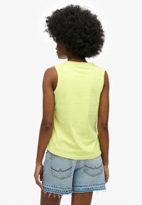 Personne aux cheveux afro bouclés portant un haut sans manches jaune clair et un short en denim bleu clair avec des ourlets effilochés, vue de dos.