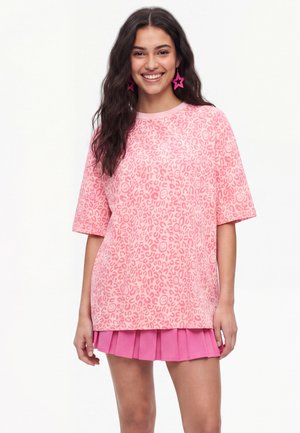 Junge Frau mit langen dunklen Haaren, die ein pink gemustertes oversized T-Shirt, einen pinken plissierten Rock und pinke sternförmige Ohrringe trägt und lächelt.