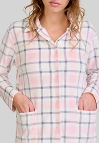 Camisa de pijama de franela con un patrón de cuadros en rosa y gris, que presenta un cierre frontal con botones y dos bolsillos laterales. Mangas largas y cuello.